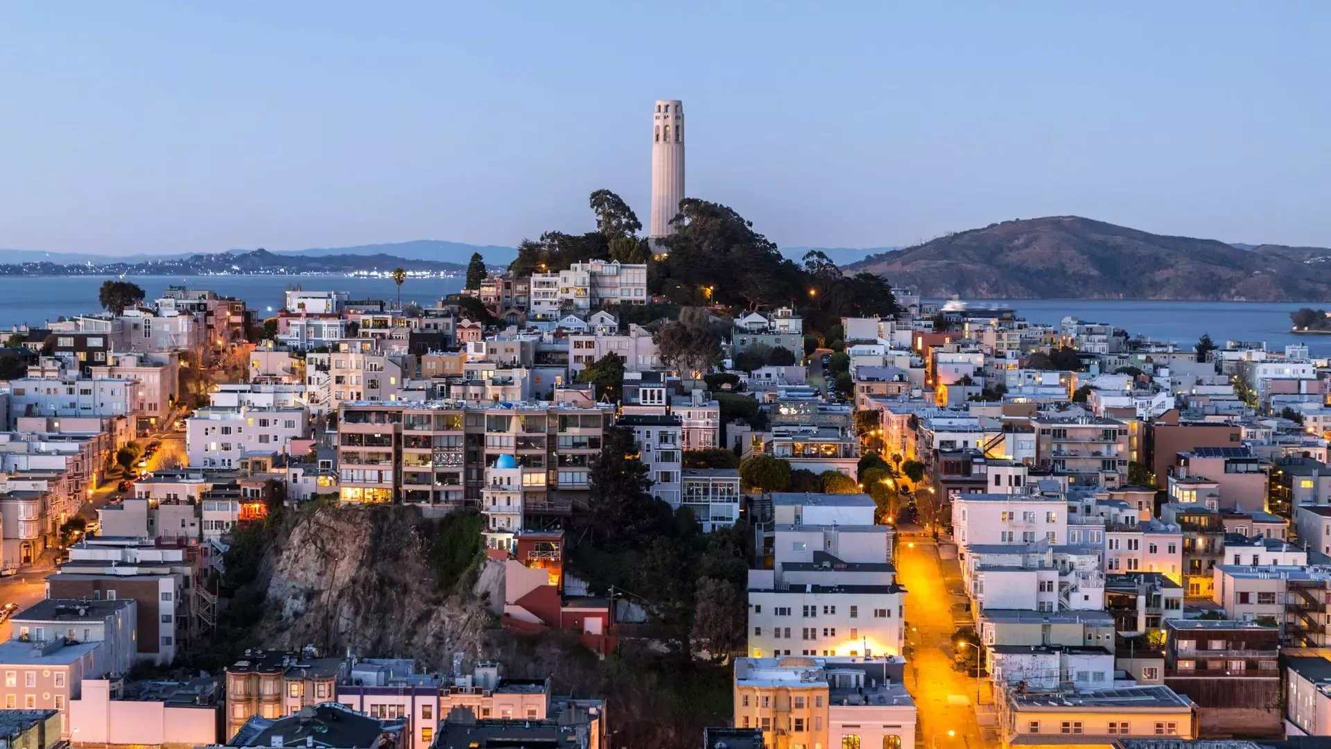 Der Coit Tower von San Francisco in der Abenddämmerung, mit beleuchteten Straßen davor und der Bucht von San Francisco dahinter.